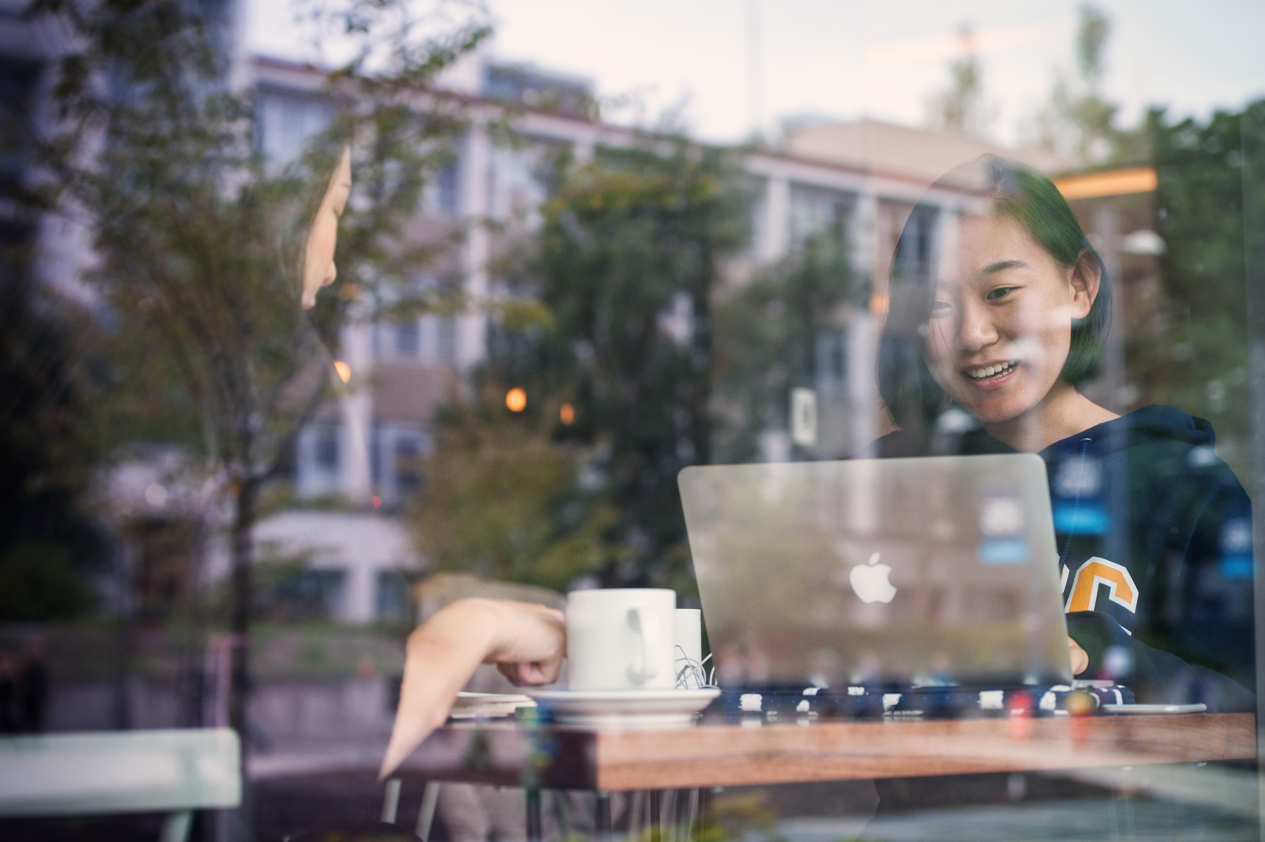 A young Asian woman smiles at her laptop in a cafe, where she sits alone at a table with a mug, earphones and a laptop bag. The large glass window reflects the outside of the cafe: trees, a building, lights.