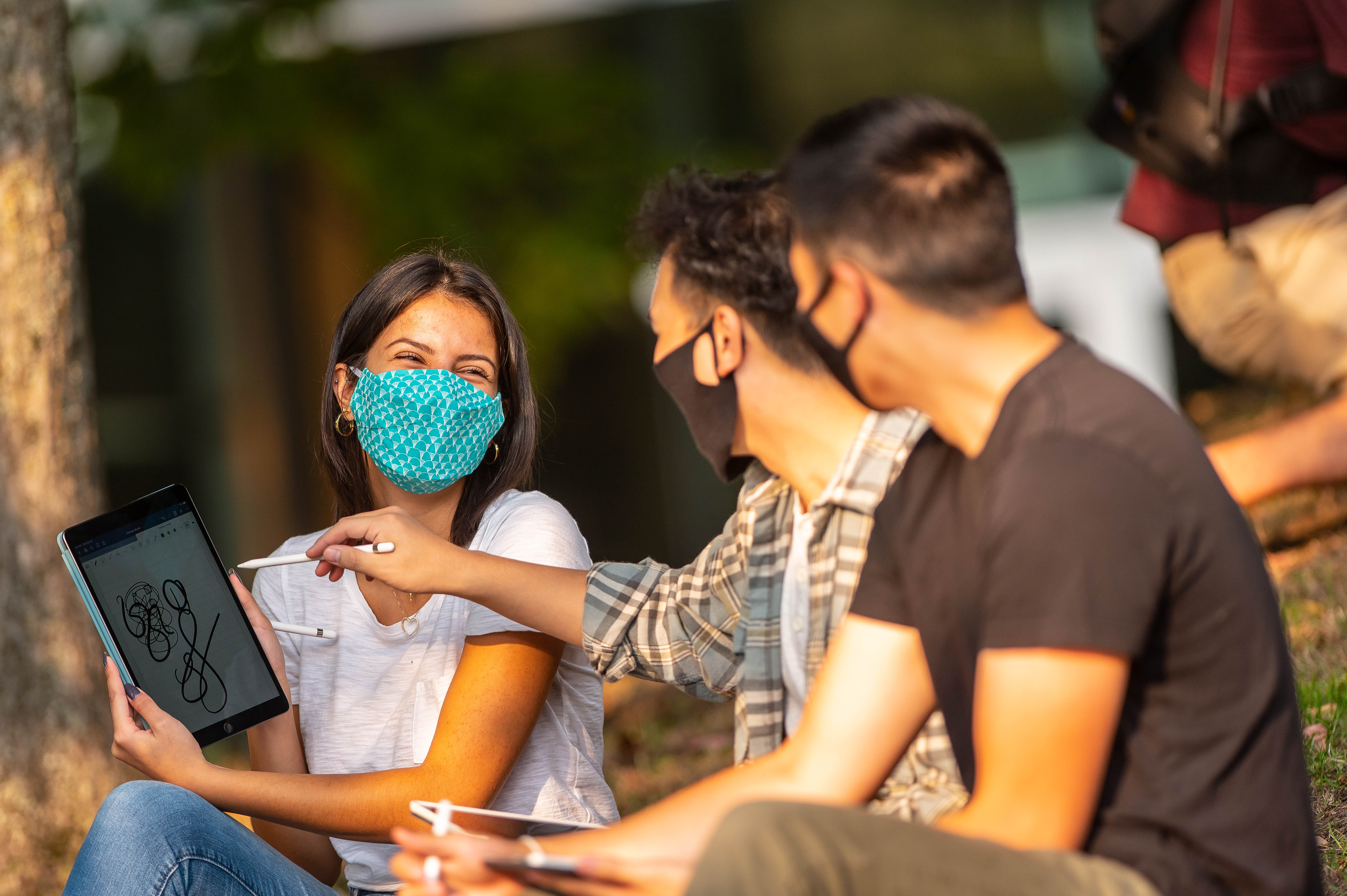 Three masked students sitting outside, on a hill. A young woman of colour smiles and holds up a tablet with a drawing of scribbles to her peers; two young Asian men who each hold styli. The one beside her holds his stylus close to her tablet, as if he is going to draw.