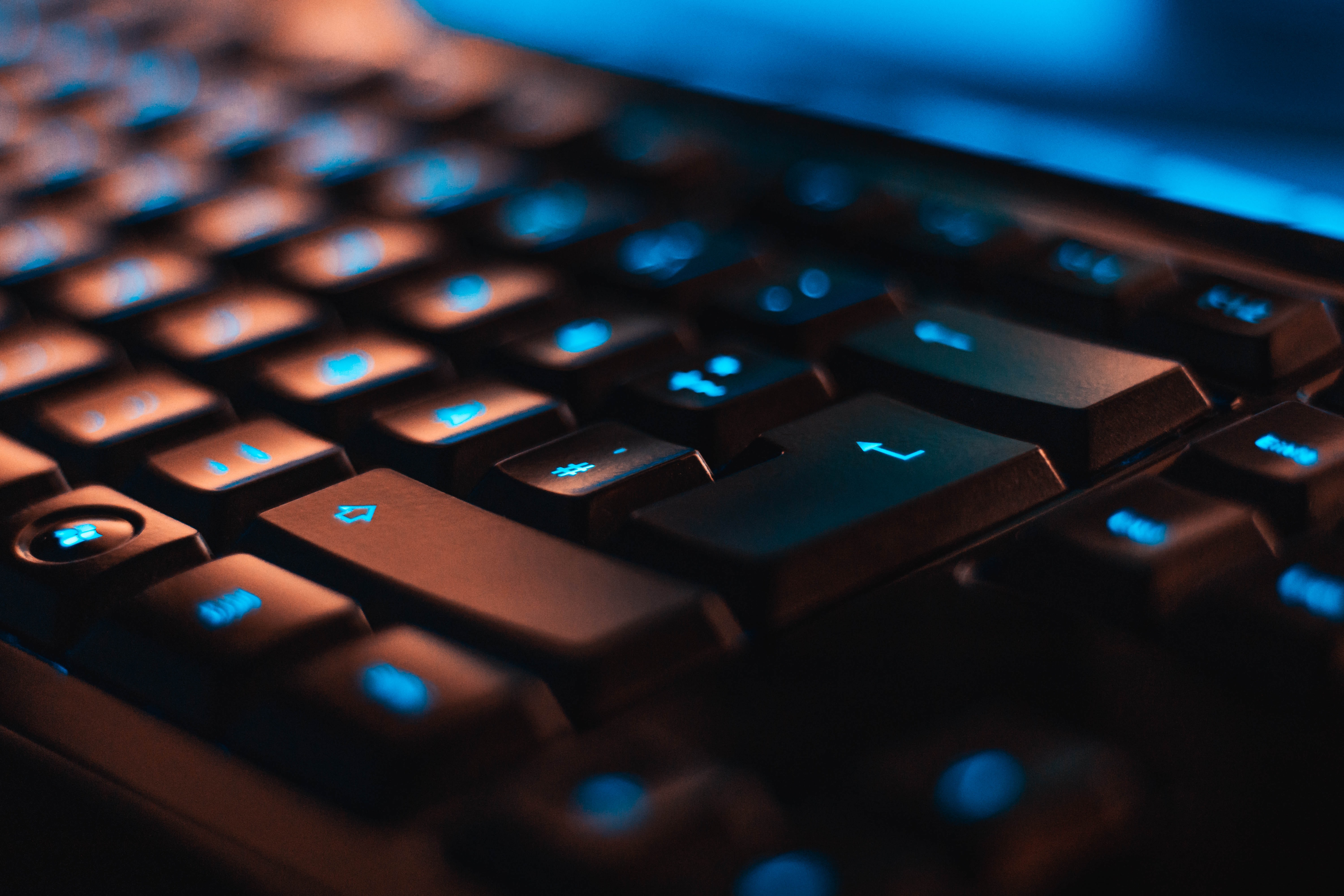 A close-up of a blue backlit keyboard with only a few keys in focus. Just above the top keys is the bottom part of the blue-lit computer screen.