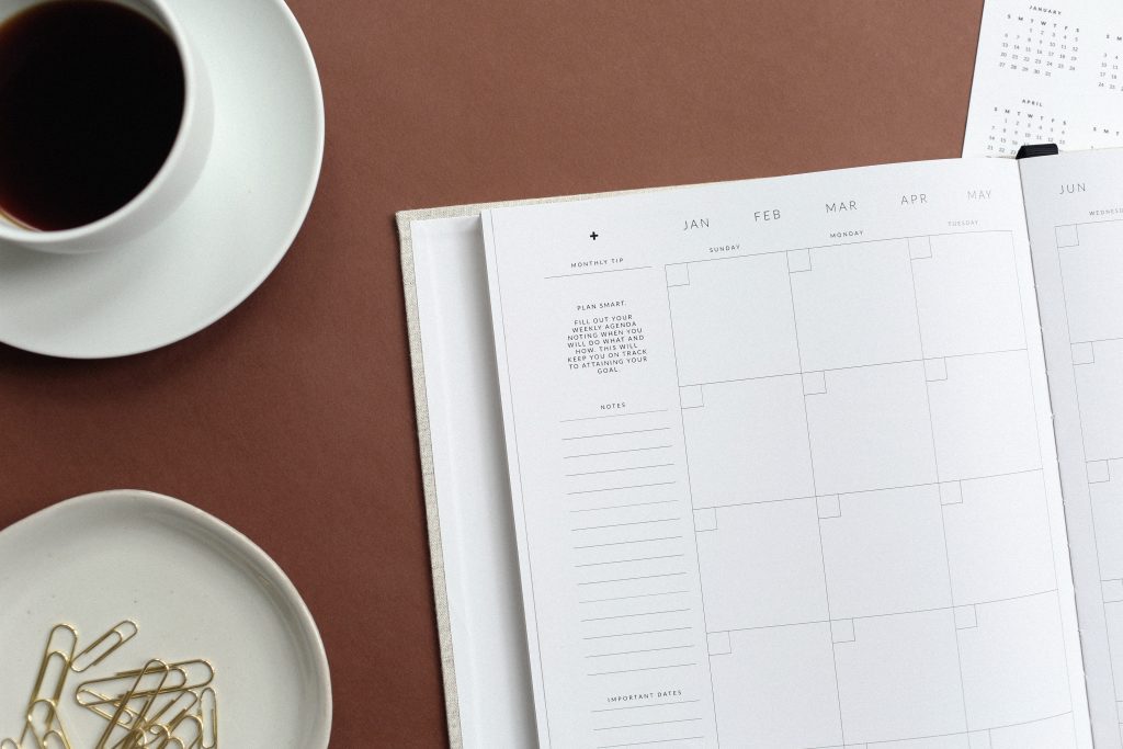 A planner, coffee cup and tray of paperclips splayed on a table. There is one corner of a paper with tiny printed calendar months.