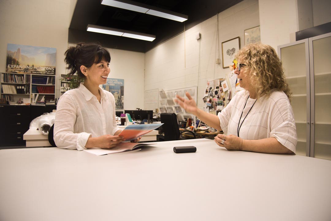 Two women (Marina de Souza and Tara Fraser) smile and talk at a table. There is a bookshelf, a bulletin board and other miscellaneous materials in the back.