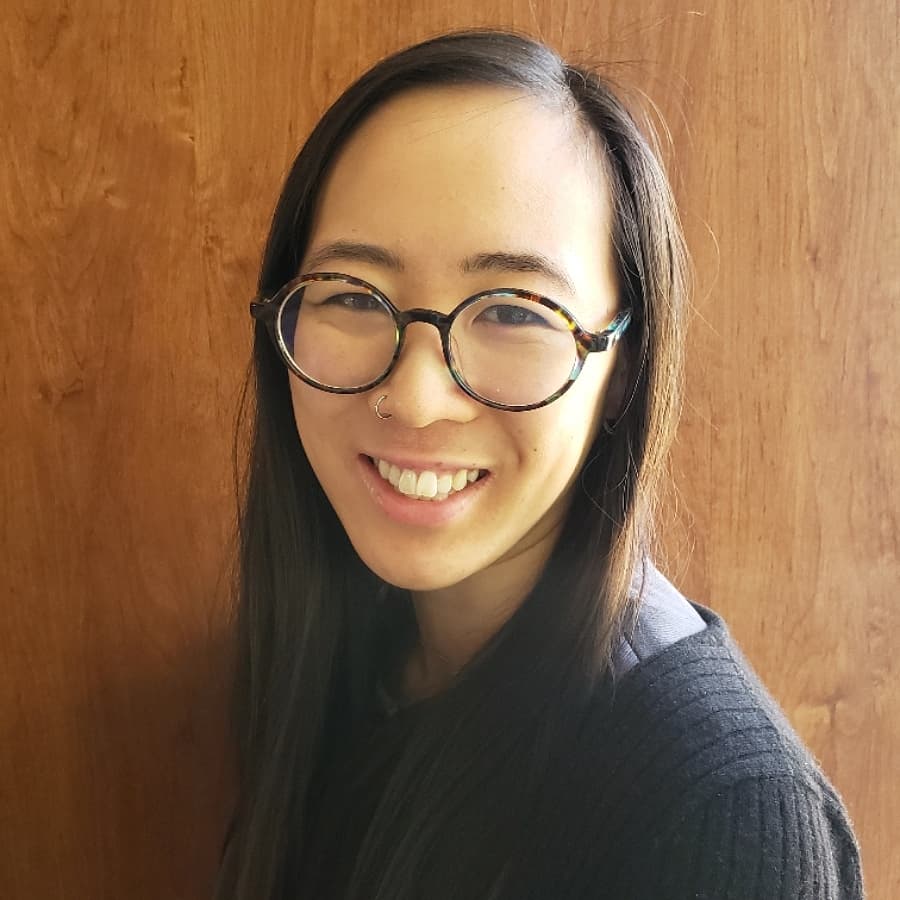 Alexandra Wong, a Chinese woman wearing glasses with long dark hair, smiles in front of a wooden background. 