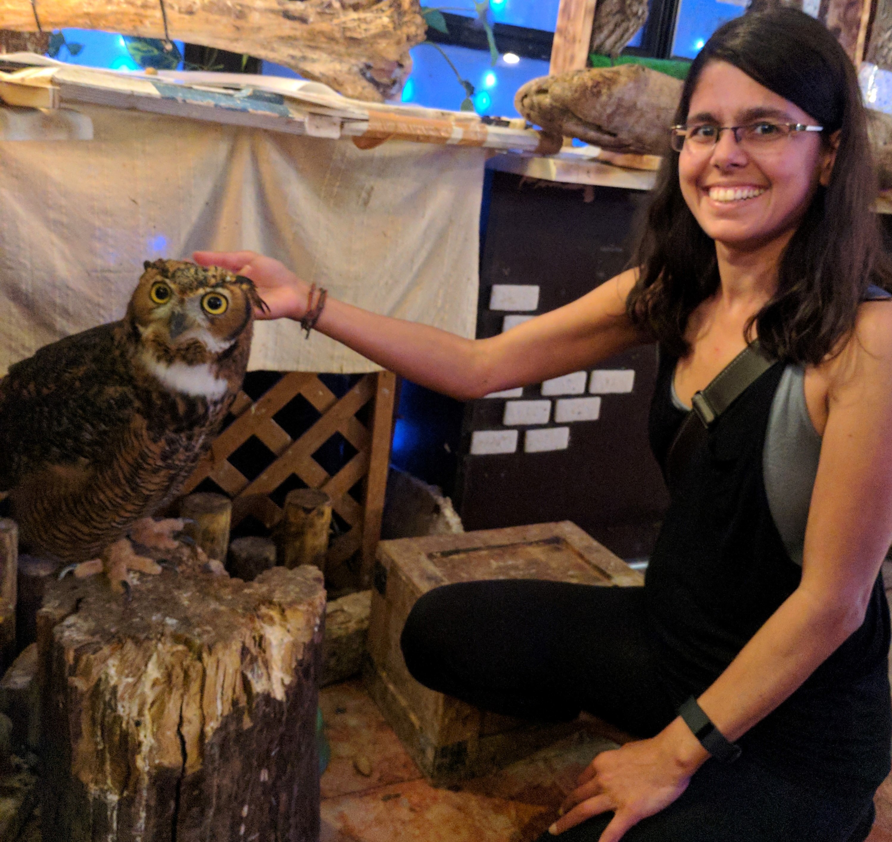 Zahra Premji crouches to the floor and smiles as she pets an owl resting on a tree stump. 