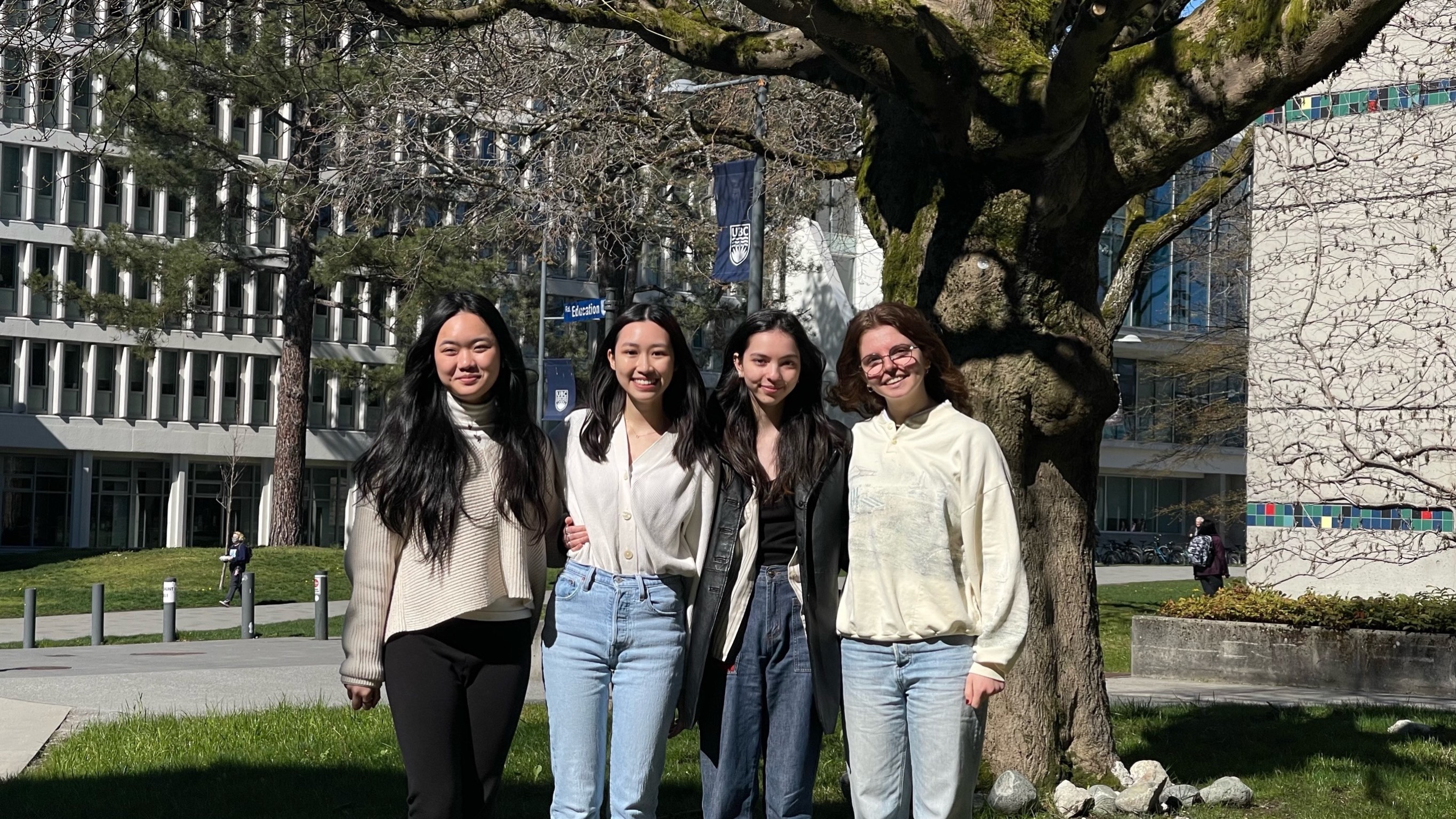 Four students smile in front of a tree (in front of buildings) on UBC Vancouver campus. 