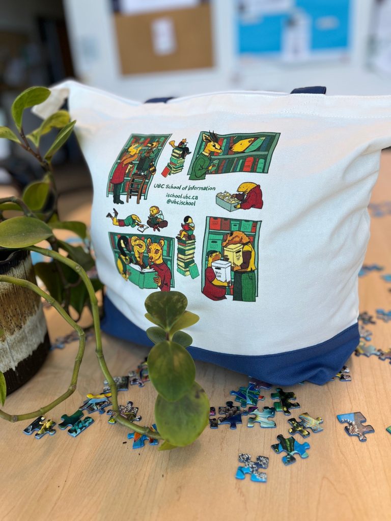 Tote bag, featuring an illustration by Maya Parker of anthropomorphized animals reading, shelving, chatting together, with the text UBC School of Information. Bag is sitting on a table amongst scattered puzzle pieces and a succulent.