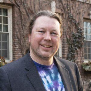 Dr Steve Easterbrook smiles at the camera, with his long straight brown hair tied back into a low ponytail. He is wearing a tie-dyed shirt in white, blue, purple, and black, with a grey blazer over it. The background of the photo is a close crop of a brick house, with vines scaling the facade.