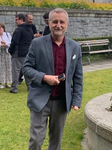 Dan has short salt and pepper hair and facial hair, and has light skin tone. He is wearing a grey suit with a burgundy shirt dress, carrying a small digital camera and smiling at the photo. He is standing casually at the U.B.C. Rose Garden.