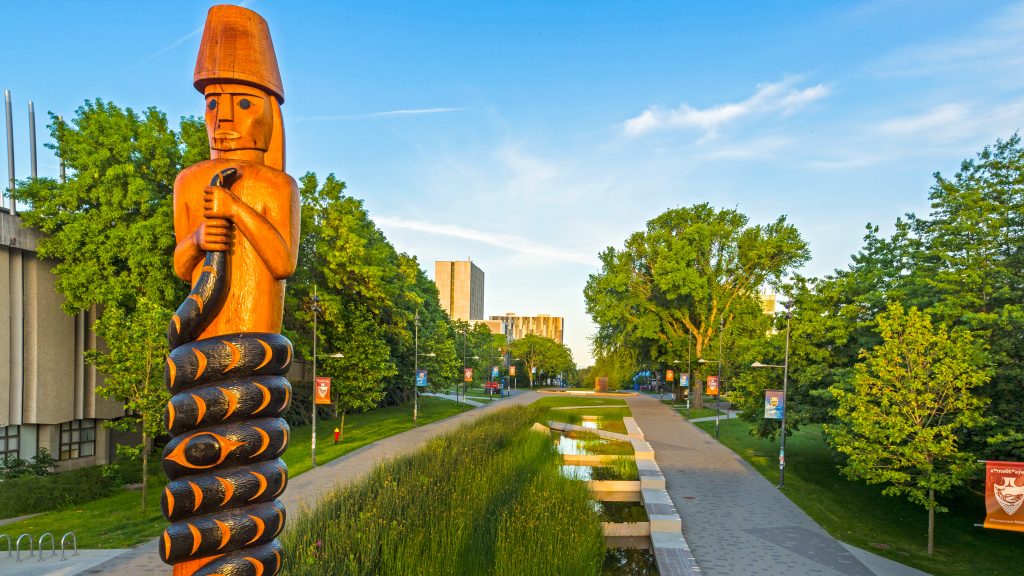 double-headed serpent post) by Brent Sparrow Jr., Musqueam. The totem pole is lit up by the golden hour sun, with a view of the U.B.C. University Boulebard behind it.