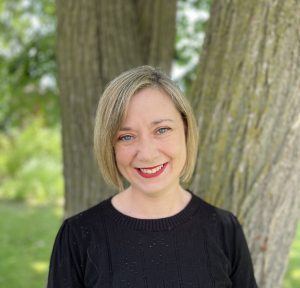 Stacy is standing in front of a tree on a sunny day, smiling.